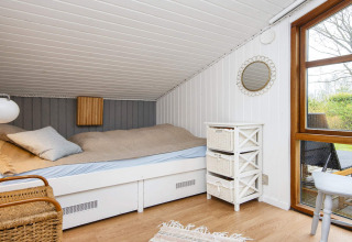 Sfeervolle slaapkamer met schuine wand, bed, witte commode met manden en tuinzicht via groot raam.
