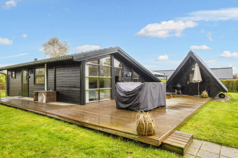 Chalet de vacances noir moderne avec grande terrasse en bois et barbecue à Hemmet, Danemark, sous ciel bleu.