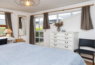 Slaapkamer in een vakantiehuis in Hemmet, Denemarken, met bed, ladekast en grote ramen met gordijnen.