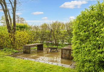 Tuin met twee banken en een vuurplaats op tegels, omringd door groen bij een vakantiehuis in Hemmet, Denemarken.