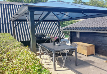 Overdekt terras met zwarte tafel, stoelen en barbecue bij vakantiewoning in Hemmet, Denemarken.