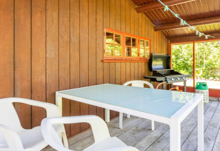 Overdekte eethoek bij vakantiehuis in Roslev, Denemarken, met glazen tafel, witte stoelen en barbecue.