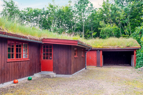 Lodge en bois rouge avec toit en herbe et garage, entouré d’arbres, près d’une maison de vacances à Roslev, Danemark.