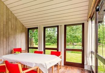 Lyst sommerhus i Roslev, Danmark, med spiseområde, røde stole og udsigt til grøn have gennem store vinduer.