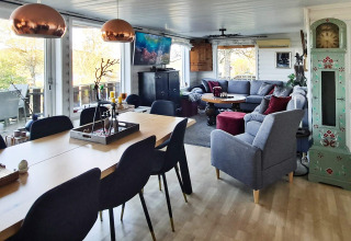 Wohnbereich einer Ferienhütte mit Esstisch, grauer Couch, vielen Fenstern und gemütlichem Ambiente.
