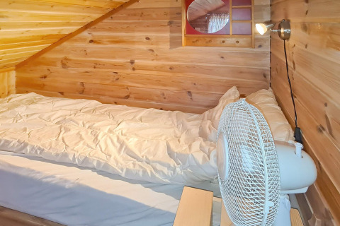 Chambre mansardée en bois avec lit simple, couette blanche, ventilateur et applique murale à Tau.