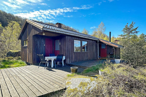 Træhytte med terrasse og havemøbler under blå himmel, idyllisk beliggende i naturskønne omgivelser.