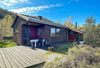Træhytte med terrasse og havemøbler under blå himmel, idyllisk beliggende i naturskønne omgivelser.