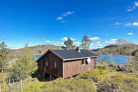 En hyggelig træhytte ved en sø, omgivet af frodig natur under en klar blå himmel og bjerge.