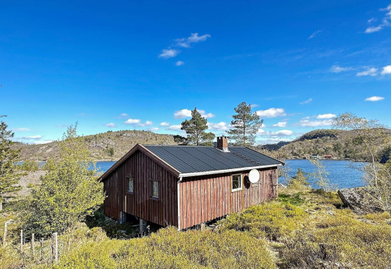 En hyggelig træhytte ved en sø, omgivet af frodig natur under en klar blå himmel og bjerge.