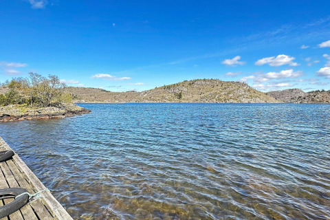 Udsigt over søen fra en træbro ved HUS 55762 HYTTE A i Danmark med bjerge og blå himmel i baggrunden.