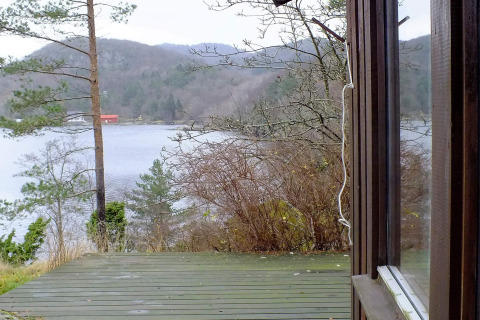 Udsigt fra HUS 55762 HYTTE A i Danmark over træterrasse, sø, bakker og en rød bygning i baggrunden.