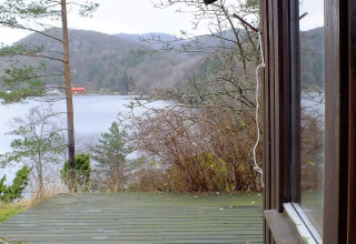 Udsigt fra HUS 55762 HYTTE A i Danmark over træterrasse, sø, bakker og en rød bygning i baggrunden.