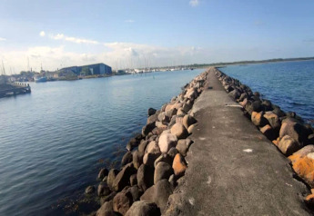 Ein steinerner Pier führt ins Meer bei Bogense, Dänemark, mit Hafen und Booten im Hintergrund bei blauem Himmel.