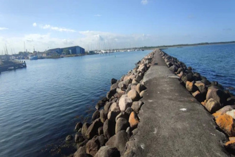 Een stenen pier loopt uit in zee bij Bogense, Denemarken, met haven en boten onder een blauwe lucht.