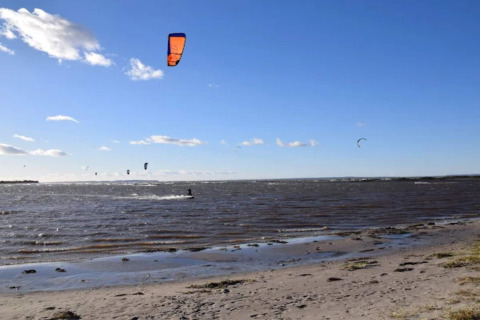 Strandscene ved Bogense, Danmark, med flere personer, der kitesurfer under en blå himmel med få skyer.