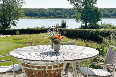 Wooden outdoor table with flower vase and chairs on patio overlooking lake and trees in Rønde, Denmark.