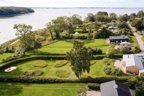 Luftfoto af feriebolig tæt ved vandet i Rønde, Danmark, omgivet af grønne områder og træer.