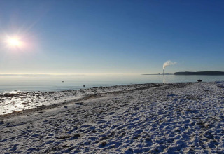 Plage enneigée et ensoleillée à Rønde, Danemark, avec fumée s’élevant d’une cheminée au loin sous ciel bleu.