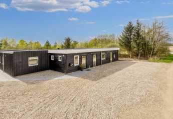 Lodge de vacances noir moderne à Øster Assels, Danemark, avec cour en gravier et arbres en arrière-plan.