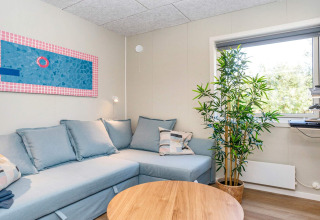 Soggiorno accogliente con divano angolare azzurro, tavolino in legno, quadro, grande finestra e pianta verde.