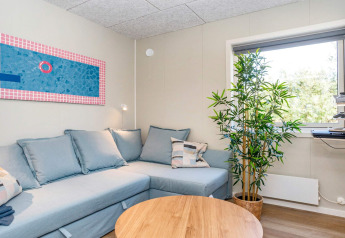 Gezellige woonkamer met lichtblauwe hoekzetel, houten salontafel, raamkunst, groot raam en groene plant.