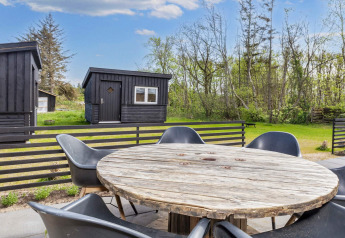 Runder Holztisch mit schwarzen Stühlen auf Terrasse einer Ferienhütte in Løgstør, Dänemark, im Grünen.