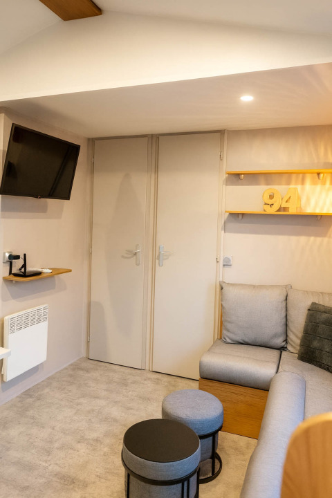 Gezellige woonkamer in vakantiewoning op Camping Papillon met TV, grijze zetel, tafeltjes en cijfer 94 op plank.