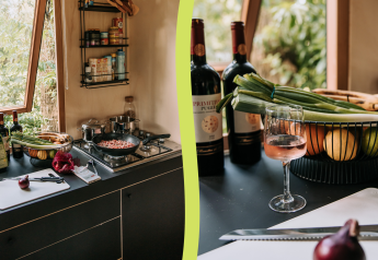 A cozy cabin kitchen with open windows, fresh vegetables, wine bottles, a cutting board, and a cooking pan.