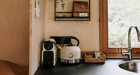 Kleine keuken in een hut met waterkoker, koffiemachine, mandje en een raam met uitzicht op groen.