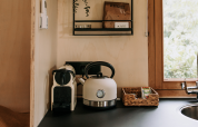 Pequeña cocina de cabaña con hervidor, cafetera, canasta y fregadero junto a la ventana con vista verde.