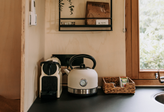 Knusse kitchenette in een hut met waterkoker, koffiemachine, mandje en raam met zicht op groen.