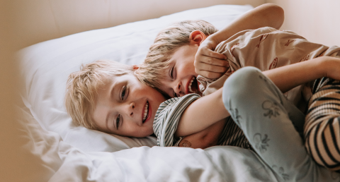 To glade børn leger og krammer på en seng i en hyggelig hytte, omgivet af blødt sengetøj og smil.