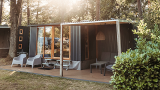 Modern cabin in the woods with a wooden deck, outdoor seating, large windows, and sunlight streaming through trees.