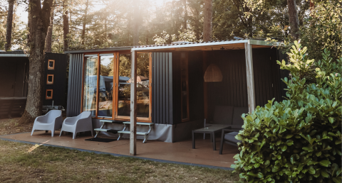 Moderne hut in een bosrijke omgeving met terras, comfortabele stoelen, grote ramen en zonlicht tussen de bomen.