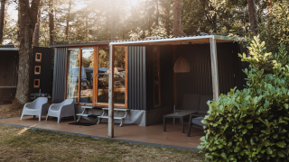 Moderne hut in een bosrijke omgeving met terras, comfortabele stoelen, grote ramen en zonlicht tussen de bomen.