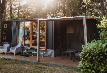 Modern cabin in the woods with a wooden deck, outdoor seating, large windows, and sunlight streaming through trees.