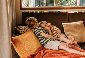 Deux enfants se détendent sur un canapé confortable dans une cabane, entourés de coussins et de lumière.