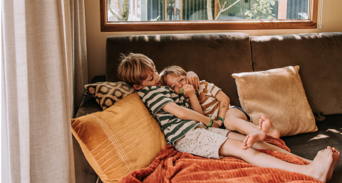 Twee kinderen knuffelen en ontspannen samen op een zetel in een gezellige cabin met kussens en licht.