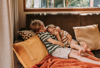 Zwei Kinder liegen entspannt auf einem Sofa in einer gemütlichen Hütte mit großen Fenstern und Kissen.