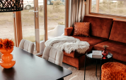 Cozy Klapkot cabin living room with a brown sofa, dining table, fuzzy throw, and large windows to nature.