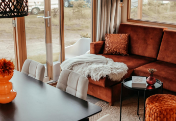 Hyggelig stue i Klapkot-hytten med brun sofa, blødt tæppe, spisebord og store vinduer til naturen.