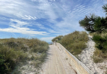 Camino de madera a través de dunas hacia la playa en Höllviken, Suecia, bajo un cielo azul con nubes.
