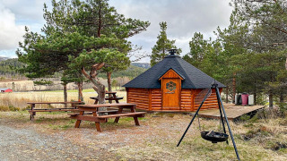 Buitenaanzicht van een ronde houten yurt met picknickbanken en vuurplek bij vakantiewoning in Lysøysundet.