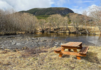 Tavolo da picnic accanto a un fiume con alberi spogli e montagne boscose sullo sfondo a Lysøysundet, Svezia.