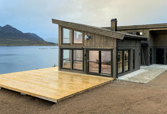 Modernes Ferienhaus in Stokmarknes mit großer Holzterrasse und Blick auf See und Berge im Hintergrund.