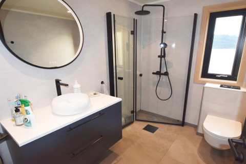 Salle de bain moderne avec douche vitrée, miroir rond, lavabo et fenêtre au gîte 5 étoiles de Stokmarknes.
