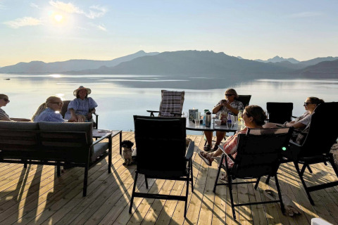 Gruppe mennesker slapper af på terrasse ved sø, bjerge i baggrunden, taget ved Stokmarknes lodge i Sverige.