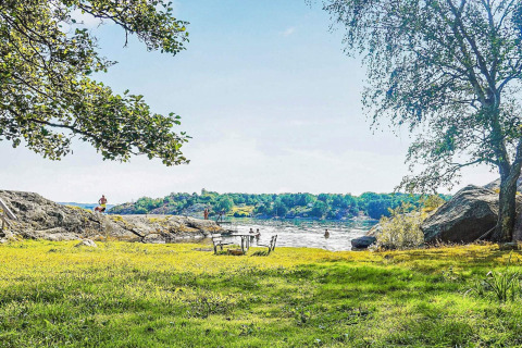 Journée ensoleillée au bord d'un lac avec rochers, arbres et pelouse près d'une cabane à Nösund, Suède.