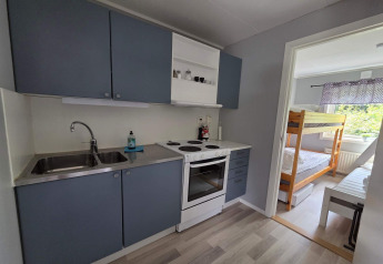 Kleine keuken met blauwe kasten en fornuis, grenzend aan kamer met stapelbed in Zweedse hut.